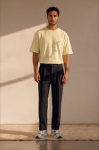 A man stands in a studio wearing a pale yellow graphic T-shirt, dark charcoal trousers, and white sneakers against a warm beige wall with soft diagonal shadows.