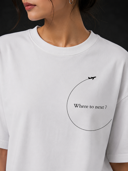 A close-up of a person wearing a white short-sleeve crewneck t-shirt with a black graphic print on the chest showing an airplane on a curved line and the text 'Where to next ?'.