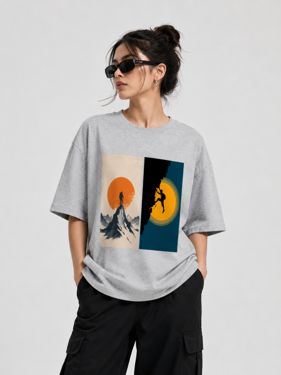 A woman modeling an oversized light gray t-shirt with a split graphic print on the front. The left side of the print shows a mountain peak under a large orange sun, and the right side shows a silhouetted figure climbing a mountain under a large yellow sun.